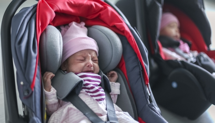 One crying newborn twin in a car seat with the other twin in the background.