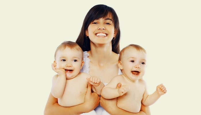 A young mother holding her twins while smiling. White background.