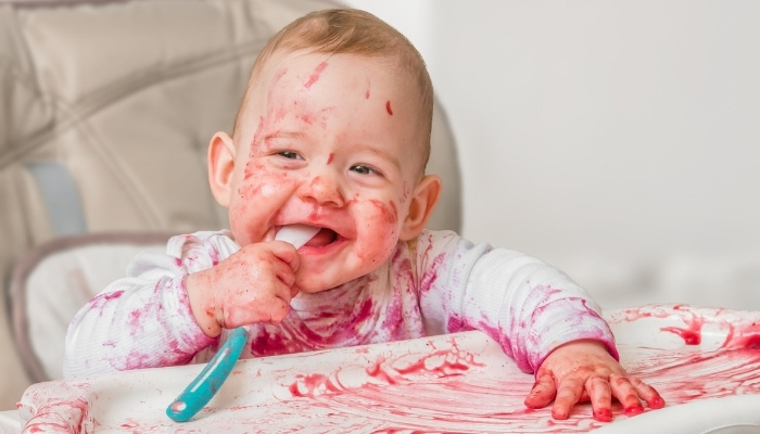A toddler in a high chair holding a spoon with a mess all over himself and the chair.