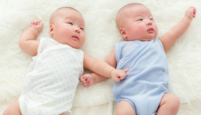 Two twin baby boys on bed with one in a blue onesie and the other in white.