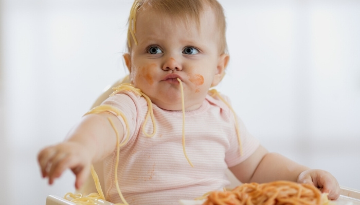 A baby in a high chair eating spaghetti and making a mess.