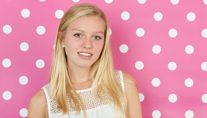 A blonde teen girl standing in front of a pink wall with white polka dots.