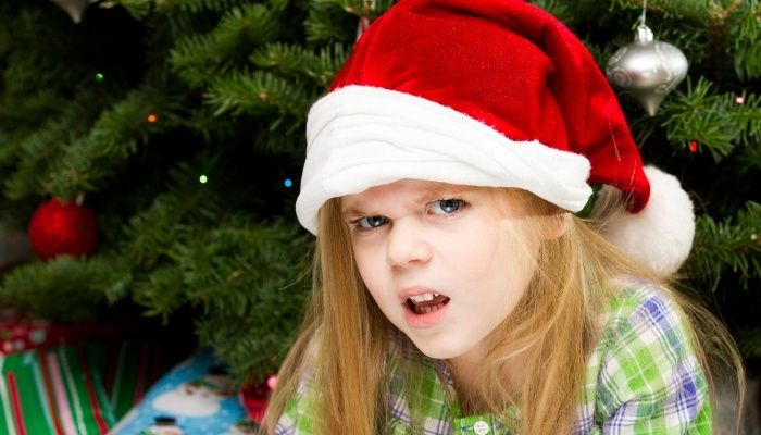 A young girl in a Santa hat sitting by a Christmas tree wearing a disgusted expression.
