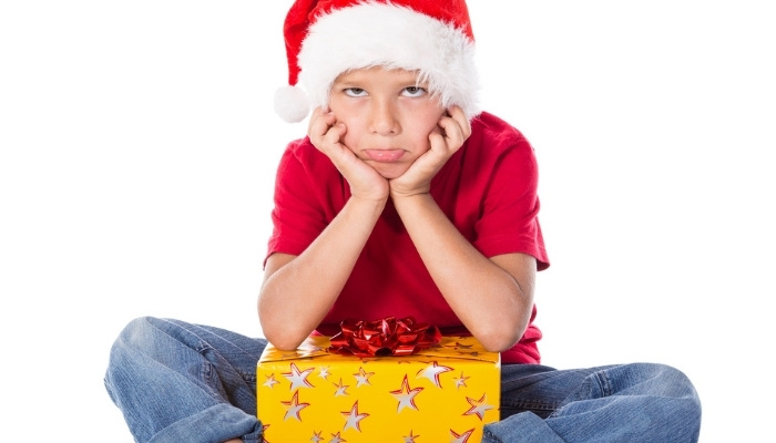 An angry boy wearing a Santa hat sitting on the floor with a gift in his lap.