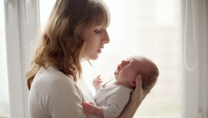 A mother holding her crying baby in front of a curtained window while she tries to soothe the child.