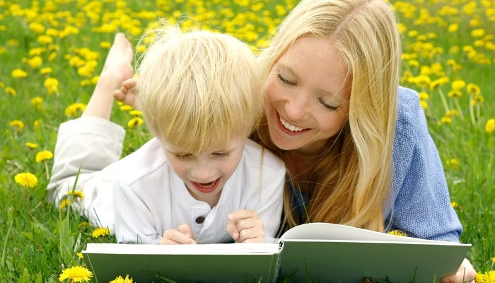 Blonde mother and son lying in grass outside while sharing a book.