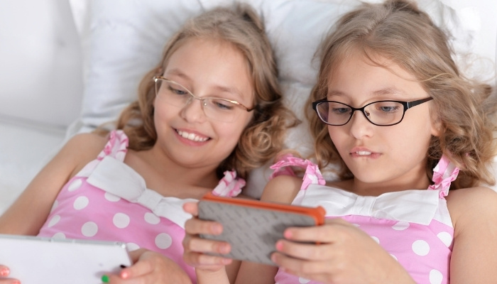 Young twin sisters wearing eye glasses relaxing in bed with their smart phones.