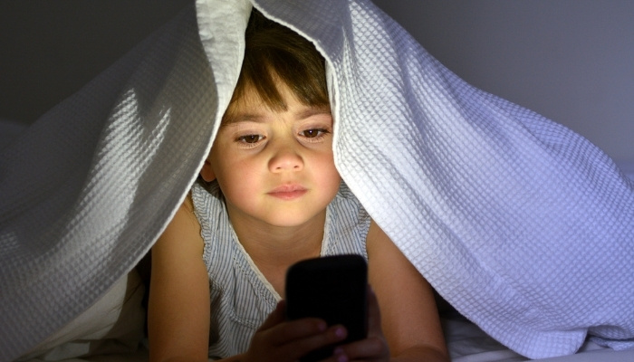 A young girl staring at a phone while under a white blanket at night.