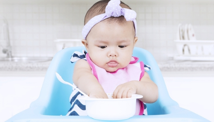 A baby girl sitting in a high chair trying to grasp food in a bowl.