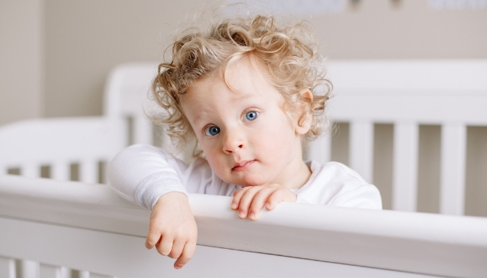 A toddler with curly blonde hair and blue eyes standing in a white crib.
