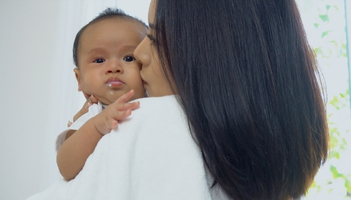 A young mother holding and kissing her baby who has some spit up on his mouth.