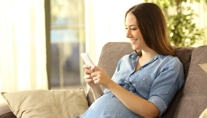 A pregnant woman looking at her smart phone and smiling while sitting on her couch.