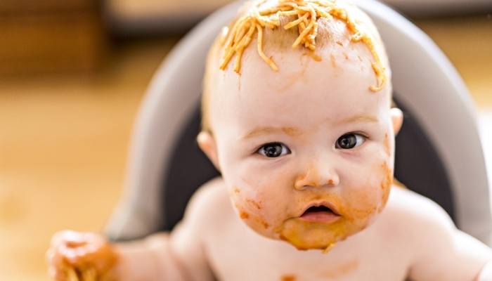 A young baby sitting in high chair messily eating spaghetti.