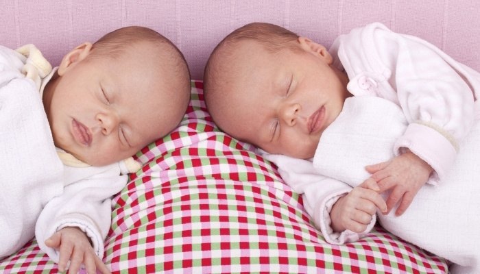 Twin babies sleeping head to head on a red-checked blanket.