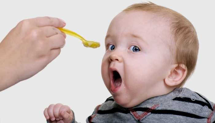A cute baby boy with his mouth open as his mother approaches with a bite of rice cereal on a yellow baby spoon.