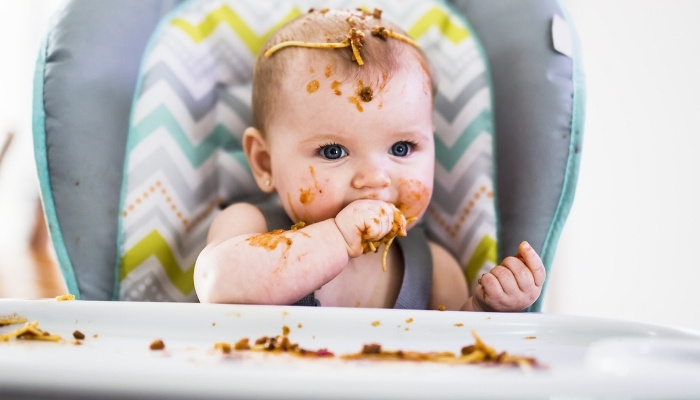 A cute baby girl sitting in a high chair trying to eat spaghetti with her hands.