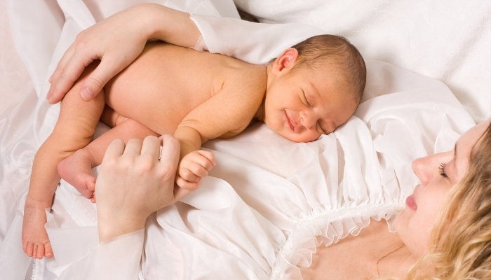 A mother holding her naked, sleeping, smiling baby on her chest.