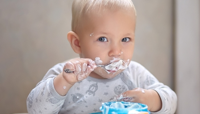 A little boy messily feeding himself yogurt from a blue bowl.