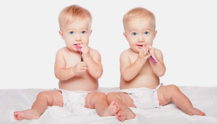 Twin brothers sitting in their diapers chewing on baby spoons against a white background.