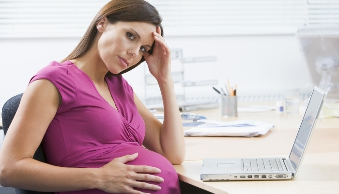 A pregnant lady in a pink shirt sitting in front of her laptop looking stressed and uncomfortable.