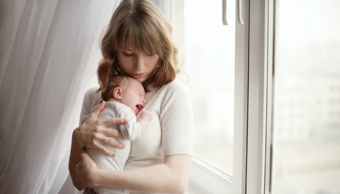 A young mother holds her crying baby near a sunny window.