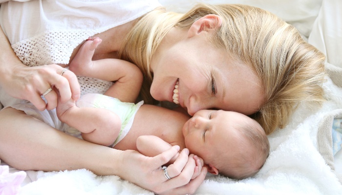 A blonde mother snuggling with her baby as they lie in bed together.