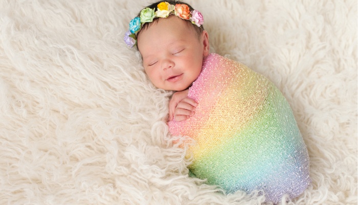 A newborn baby girl wearing a rainbow swaddle and matching headband.