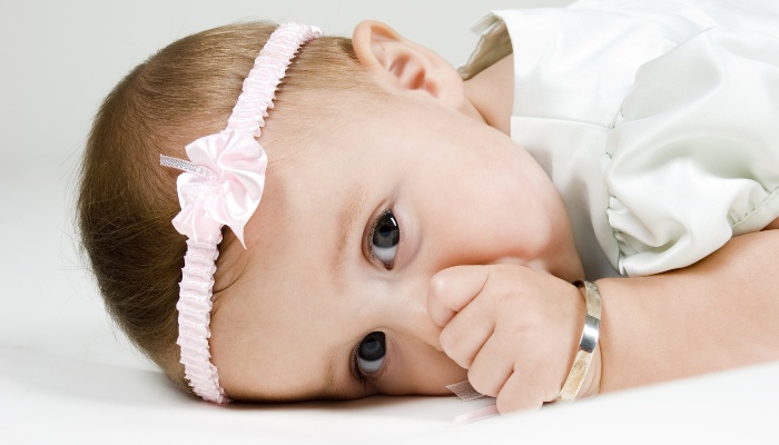 A baby girl with a headband and bracelet lies on her tummy while sucking her thumb.
