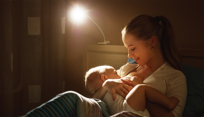 A young mother breastfeeds her baby at nighttime with a small lamp glowing in the background.