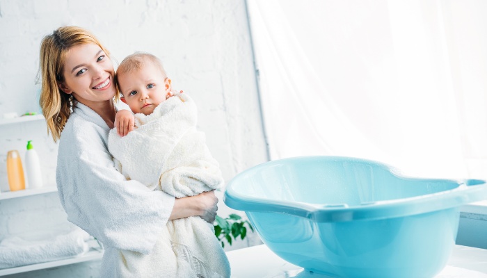A mother holding her baby wrapped in a bath towel standing near a baby bathtub.