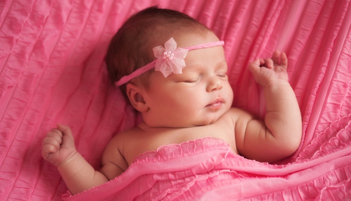 A newborn baby girl wearing a pink headband and lying on pink sheets under a pink blanket.