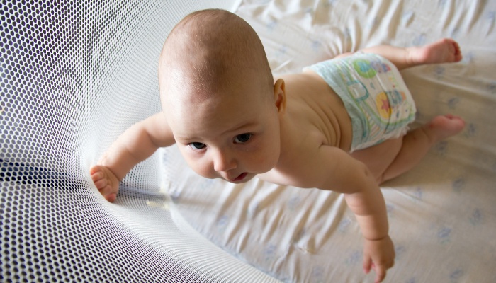 A baby wearing only a diaper leaning against the side of his playpen.