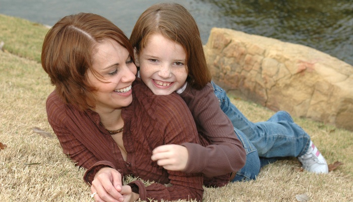 A happy girl lies on her smiling mother's back while they spend time at a pond.