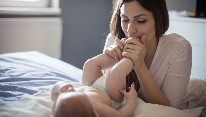 A young mother kissing her baby's feet while he lies on the bed.