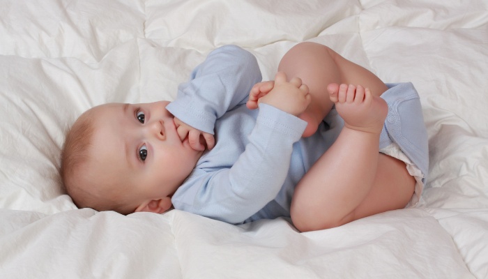 A cute baby boy on his back holding his foot and sucking on his fingers and bottom lip.